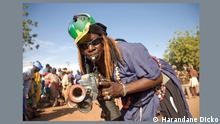 Ein afrikanischer Mann hat eine Trillerpfeife in der Hand und guckt in die Kamera.