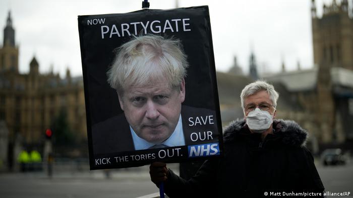 A masked protester stands in front of the Houses of Parliament holding an anti-Johnson placard reading 'Now Partygate. Kick the Tories out. Save our NHS' 