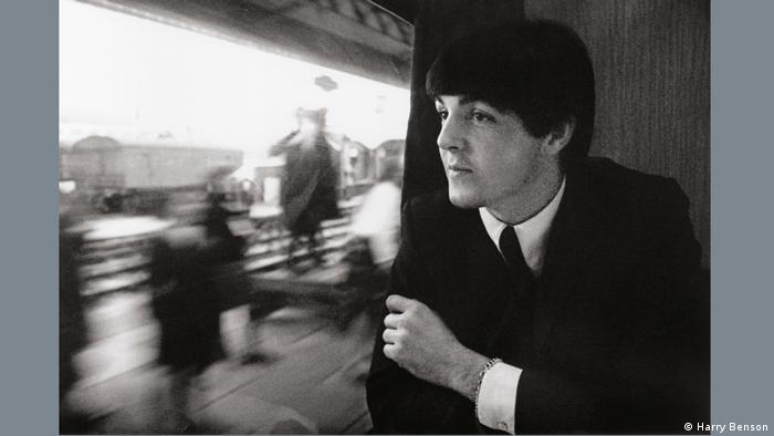Paul McCartney sitting by a train's window., wearing a tie and suit. Blurry people can be seen on the platform.