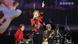 Mick Jagger stands on stage and sings with one hand pointing in the air while wearing a red and black striped jacket and dark jeans. Mick Jagger stands on stage and sings with one hand pointing in the air while wearing a red and black striped jacket and dark jeans.