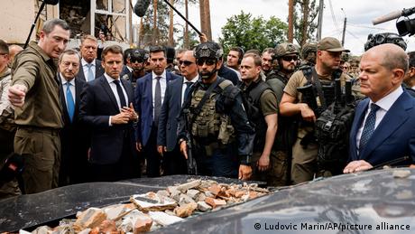 Zu sehen sind unter anderem der französische Präsident Emmanuel Macron und der deutsche Bundeskanzler Olaf Scholz, die Irpin in der Ukraine einen Besuch abstatten, um ihren Beistand zu demonstrieren. Sie werden von Soldaten begleitet und schauen auf ein zertrümmertes Auto.