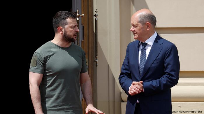 Ein ernst dreinblickender Mann mit Bart im olivfarbenem T-Shirt und ein lächelnder Mann mit Halbglatze im Anzug stehen sich in der Sonne gegenüber