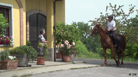 A man rides a horse and approaches the entrance to a house, as an older woman waits in the doorway