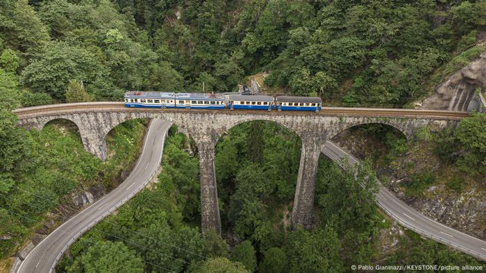 A small train crosses a viaduct, below one can see roads and forest