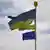 A tattered Ukrainian flag flies next to an EU flag against a partly cloudy sky