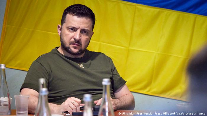 Ukrainian President Volodymyr Zelenskyy, wearing an army-green T-shirt, sits in front of the Ukrainian flag at a table, looking concerned