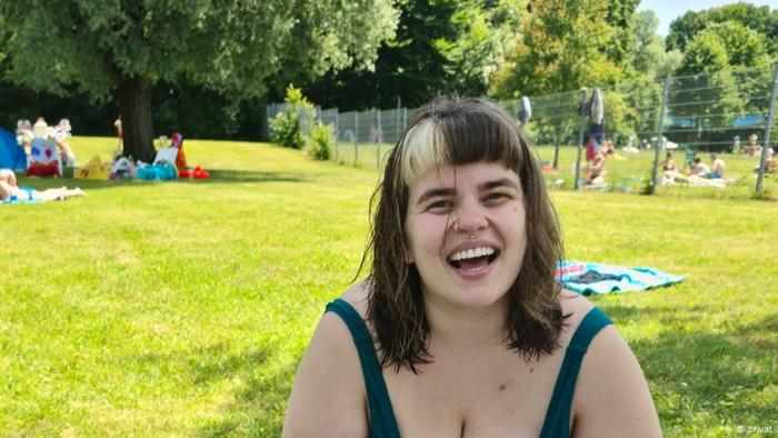Una mujer joven con cabello oscuro hasta los hombros y una racha decolorada se sienta en un campo de hierba, sonriendo
