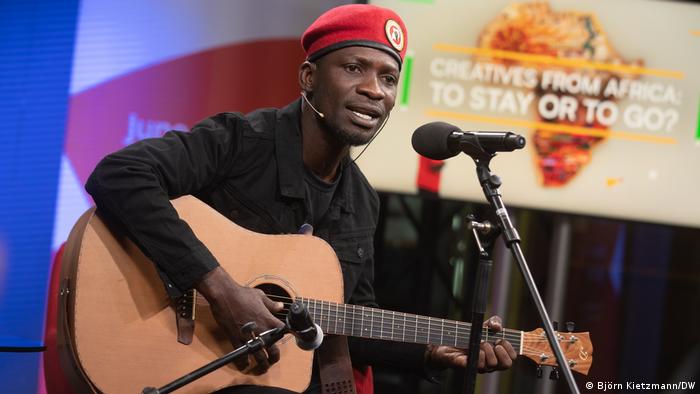 Bobi Wine in black shirt and red hat singing and playing the guitar