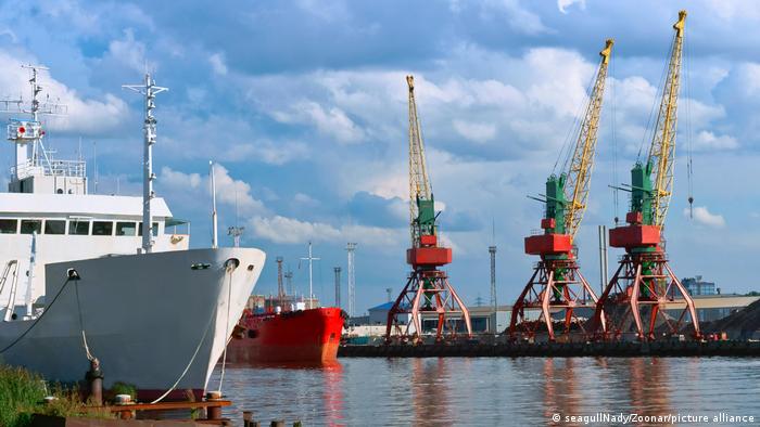 A white ship and a red ship at anchor in a port, three cranes and a blue sky in the background