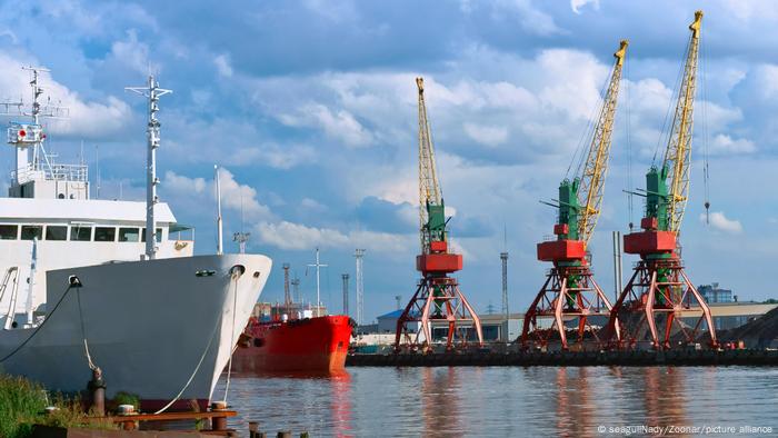 A white ship and a red ship at anchor in a port, three cranes and blue sky in the background