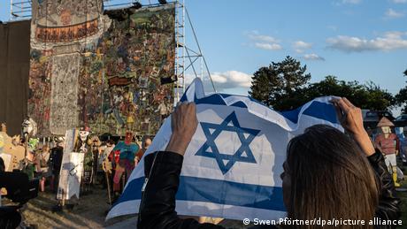 A woman holds the flag of Israel in front of a huge artwork on scaffolding.