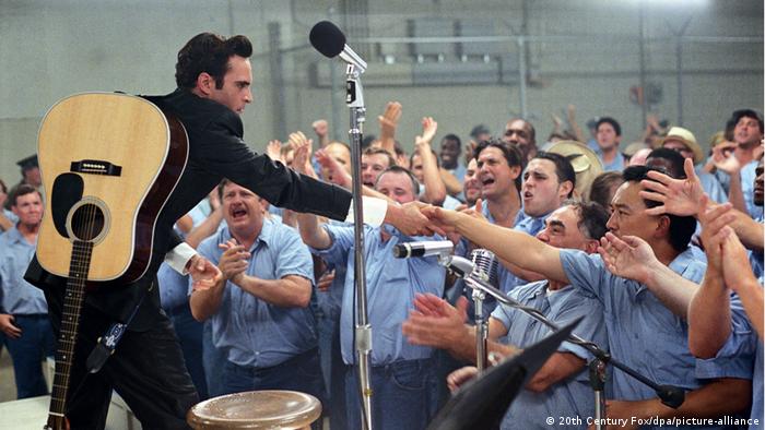 Joaquin Phoenix as Johnny Cash, man stands in front of a crowd of men dressed in blue shirts, stretches out a hand to one of them 