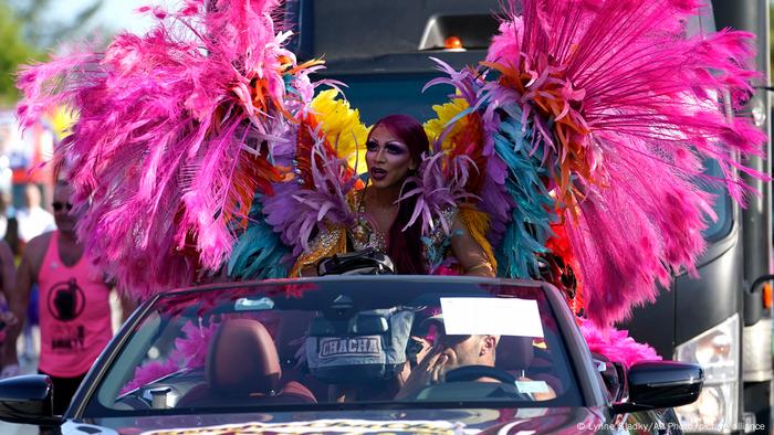 Eine Drag Queen mit regenbogenfarbenem Gefieder steht während einer Parade in einem offenen Cabriolet.