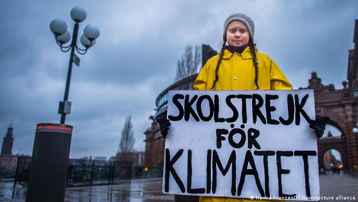 A young girl dressed in a yellow raincoat and with a beanie on her head holds up a placard on which is written in Swedish School strike for the climate.