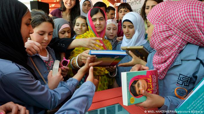 Malala Yousafzai distributing copies of her book I am Malala to a group of women surrounding her.