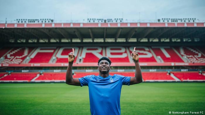 Taiwo Awoniyi stands on a football pitch, holding up both hands and pointing to the sky