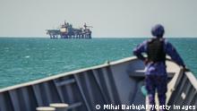 A soldier in the prow of a small boat, out of focus, gazes across a turquoise sea at an oil platform in the distance