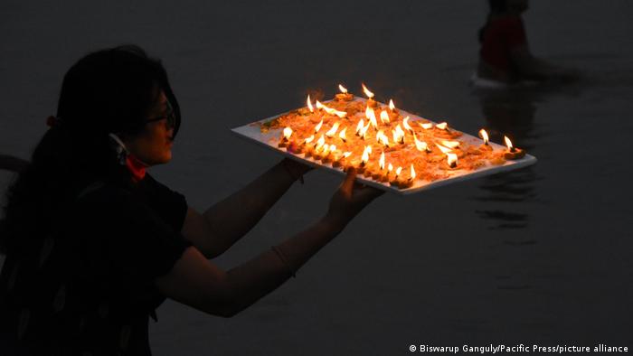 A woman holds a tray with lit candles and flowers in her hands and stands in front of the river Ganges at night
