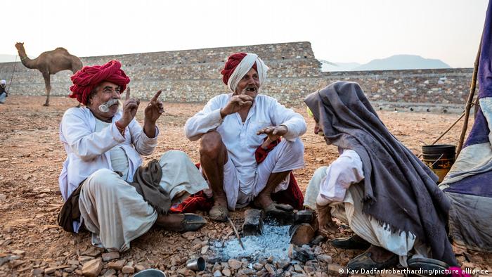 Three men with cloths on their head are sitting together in front of an extinguished fire, gesticulating and laughing. In the background is a wall, a camel and mountains.