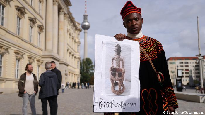 A man in a red hat holds up a poster featuring a statue that reads #bringbackngonnso