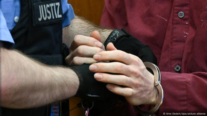 Blick auf Hände in dunklen Handschuhen von einer Person in einer Weste mit der Aufschrift Justiz, die einer anderen Person im roten Hemd die Handschellen löst Blick auf Hände in dunklen Handschuhen von einer Person in einer Weste mit der Aufschrift Justiz, die einer anderen Person im roten Hemd die Handschellen löst