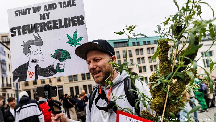 Un demostrador que sostiene una planta de cáñamo, otro que sostiene un cartel que dice: cállate y toma el dinero de mis impuestos.
