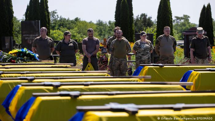 Särge bedeckt mit der ukrainischen Flagge und einem Kreuz. Dahinter Soldaten.