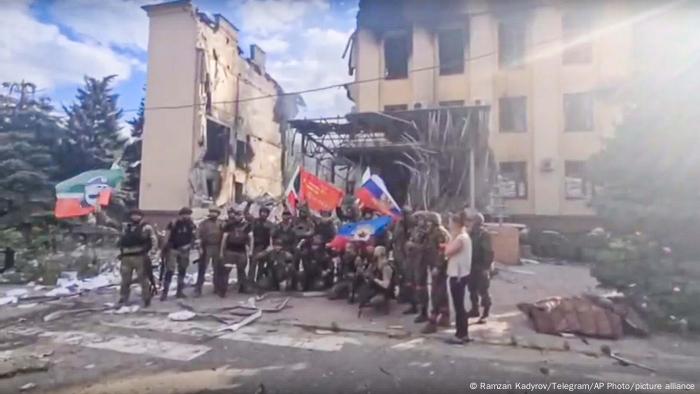 Esta foto tomada del vídeo proporcionado por el canal oficial de Telegram de Ramzan Kadyrov publicado el sábado 2 de julio de 2022 muestra a las tropas rusas, incluidos los soldados del regimiento checheno, ondeando las banderas nacionales de Rusia y de la república chechena mientras posan para una foto frente a un edificio destruido en Lisichansk.