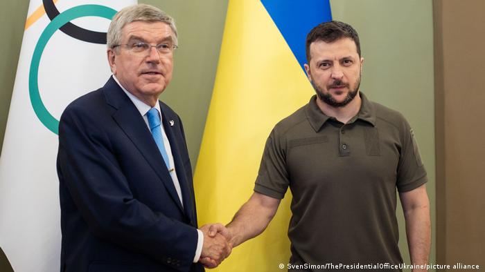 Volodymyr Zelenskyy (right) shakes hands with IOC President Thomas Bach in front of a Ukraine flag