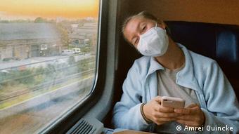 Amelie Quincke, blond im hellblauen Fleecepulli und mit weißer Maske hält ihr Handy in der Hand. Sie sitzt in einem fahrenden Zug.
