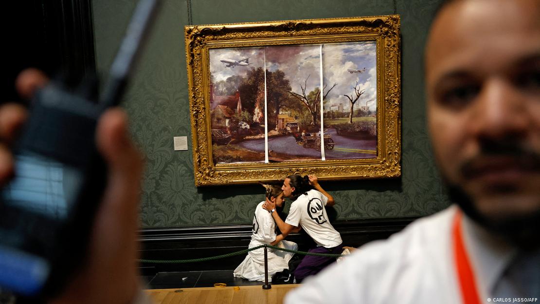 Em primeiro plano, desfocado, um segurança de museu. Ao fundo, um homem beija a testa de uma mulher. Eles estão em frente a uma obra de arte. Sobre a pintura original, foram colados papeis com uma nova imagem.