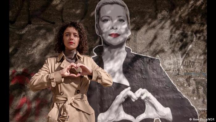 Жінка в коричневому пальті власноруч малює знак серця, репродукції вуличного мистецтва на стіні за нею, де також зображена жінка, що робить такий самий знак серця 