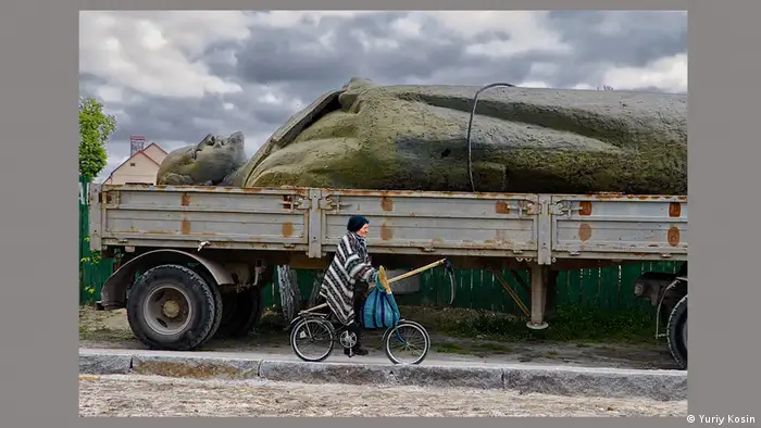 Eine gestürzte Lenin-Statue liegt auf der Ladefläche eines Lastwagens. Eine Bäuerin fährt auf ihrem Fahrrad vorbei, sie trägt eine Hacke und eine Tasche mit frisch geernteten Früchten.