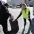 Auf einem gepflasterten Platz hilft eine junge Frau mit signalfarbener Schutzweste einer älteren Frau mit ihrem Koffer