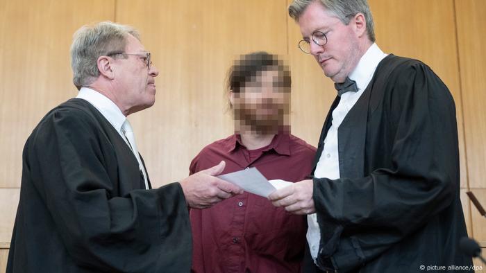 Rechts und links zwei Männer in weißen Hemden und dem schwarzen Umhang von Anwälten, in ihrer Mitte steht ein Mann im roten Hemd mit Vollbart, sein Gesicht ist verpixelt Rechts und links zwei Männer in weißen Hemden und dem schwarzen Umhang von Anwälten, in ihrer Mitte steht ein Mann im roten Hemd mit Vollbart, sein Gesicht ist verpixelt