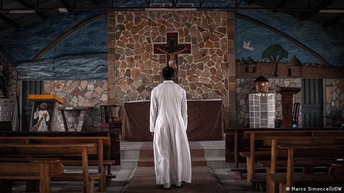 Father Igor Armand Kassah, dressed in a white cassock, stands in front of a church altar 