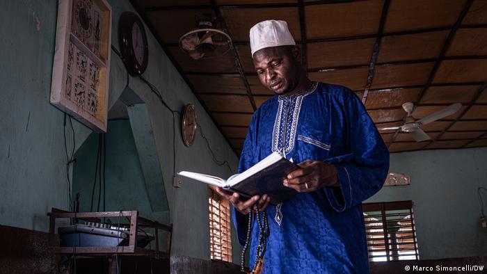 Mounou Y'Moussa, the imam of Porga's central mosque, wearing a bright blue thawb, stands reading the Quran 