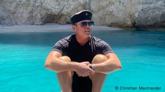Christian Mastrander, Eigentümer von Cotton Club Zakynthos mit Mütze, Sonnenbrille und in Shorts vor dem Meer