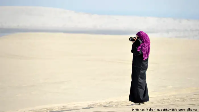A female photographer wearing a black abaya and a headscarf stands at a beach in Qatar 