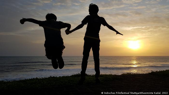 In a still from 'Kasiminte Kadal' two people holding hands and looking at the sea at sunset, one of them is jumping.