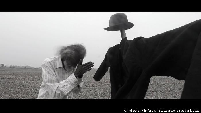 Still from 'Adieu Goddard': black-and-white photograph of a man in a prayer position while looking at a scarecrow made of a hat and coat.