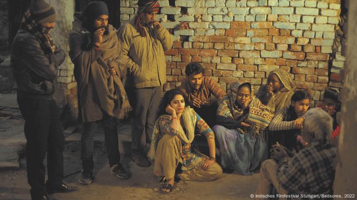 Photo du film « escarres » : un groupe de personnes en costume traditionnel, trois hommes sont debout et fument et les autres sont assis contre un mur.