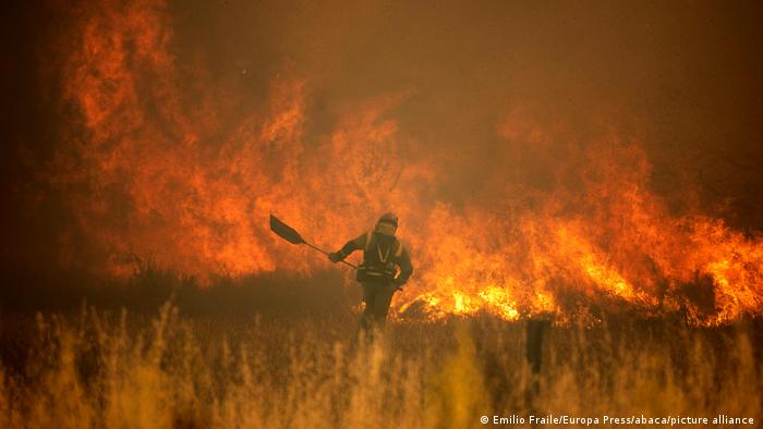 Ein Feuerwehrmann mit einer Schaufel vor einem Meer aus Flammen in einer Graslandschaft