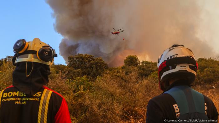 Two firefighters look on as a helicopter flies to drop water on a forest fire
