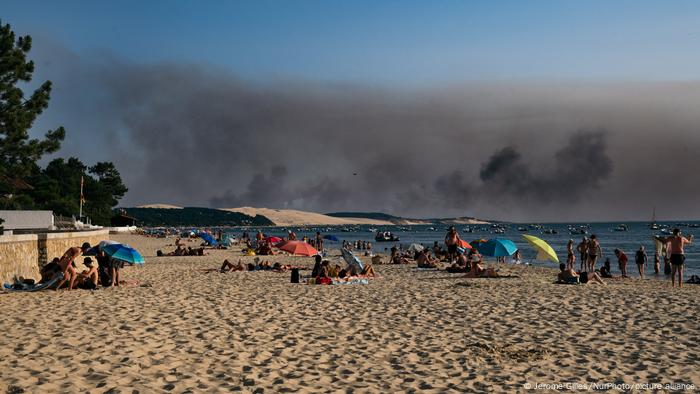 People sit on a beach on France's Atlantic coast with black smoke from a forest fire visible in the background