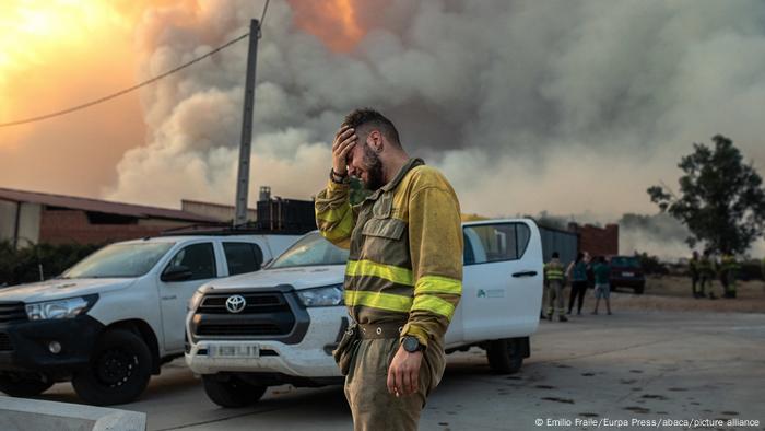 Un bombero en ropa de trabajo, visiblemente agotado y con cara de sufrimiento, se sujeta la frente con la mano. Al fondo se ven las torres de humo por los incendios.