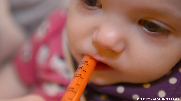  Ein Kleinkind hat eine orangefarbene Spritze im Mund, wie sie Packungen mit Fiebersäften beiliegt
