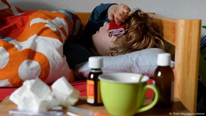 Deutschland | Medikamente und Apotheken | Fiebersaft. Ein Kind liegt im Bett und hat eine Kompresse auf der Stirn. Im Vordergrund stehen auf einem Tisch eine grüne Tasse und zwei Flaschen mit Medikamenten. Davon eine mit Fiebersaft