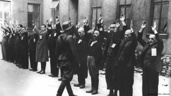 A line of mostly middle-aged and older men in dark suits stand in the snow in front of a building with their hands raised, a soldier with a round helmet right in front of them. The photo is black and white.