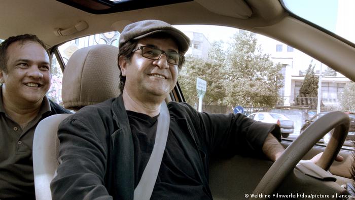 Filmstill aus dem Film Taxi Teheran. Der Regisseur sitzt selbst am Steuer, ein Fahrgast hinten.